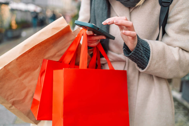 Cropped image of female hands holding red holiday gifts packages used phone on the city street near the shopping center. Retail sale concept. Shopper. Sales. Shopping Center.Cyber Monday.Black Friday - Photo, image