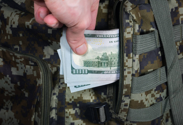 Man is putting in a military backpack dollars. Dark green Camouflage khaki bag with money. Migrants and Refugees. Looting and Robbery concept. - Photo, Image