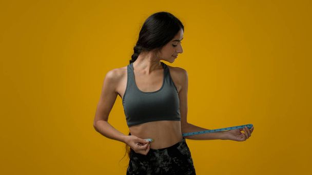 Stüdyo sarı arka planda mutlu bir kadın sporcu bayan sporcu bayan bel ölçüsünde vücut şekli ve bant kontrolü diyet sporu diyeti zayıflama sonucu zayıflama - Fotoğraf, Görsel