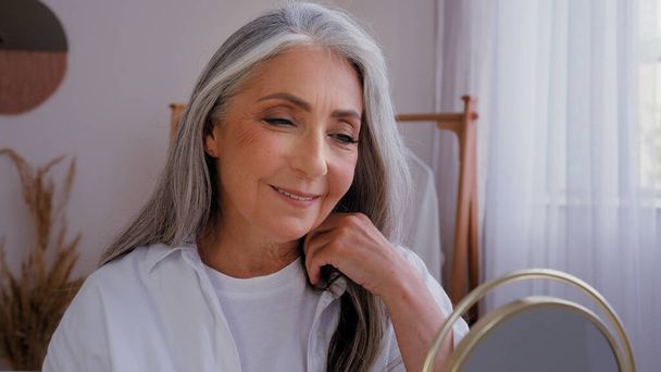 50-es évek yeas idős hölgy ősz hajjal élvezi a bőr nedvességét nézi tükör reflexió érintés arc krém alkalmazása 60-as évek idősebb érett nő ellenőrizze kozmetikai eredmények arc kozmetikumok megható arc - Fotó, kép