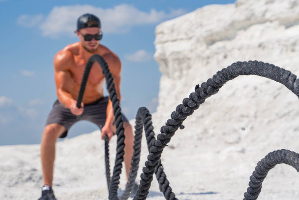 A strong man in a baseball cap and black glasses is training with ropes in nature - 写真・画像
