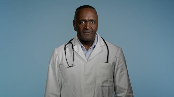 Зрелый уверенный в себе счастливый доктор, стоящий на сером фоне студии со скрещенными руками, радостно улыбающийся афроамериканец, врач-терапевт в медицинском халате, позирующий в помещении, глядя в камеру - Фото, изображение