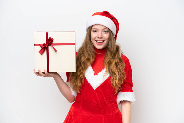 Молодая белая женщина в рождественском платье с подарком на белом фоне с сюрпризом и шокированным выражением лица - Фото, изображение