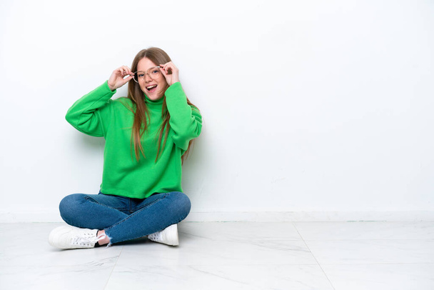 Young caucasian woman sitting on the floor isolated on white background with glasses and surprised - Photo, Image