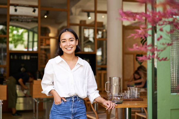 Portrait of young businesswoman in her own cafe, manager standing near entrance and inviting you, posing in white plain shirt and jeans. - Photo, Image