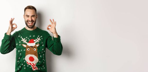 Різдво, свята та святкування. Задоволений красивий хлопець в светрі, показуючи хороші знаки і посміхаючись, хвалити щось чудове, стоячи на білому тлі
. - Фото, зображення
