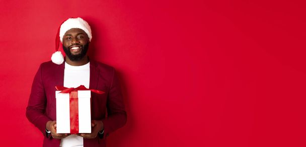 Boże Narodzenie, Nowy Rok i koncepcja zakupów. Szczęśliwy brodaty Afroamerykanin trzyma obecny Boże Narodzenie, uśmiecha się do kamery, stoi w kapeluszu Mikołaja na czerwonym tle. - Zdjęcie, obraz