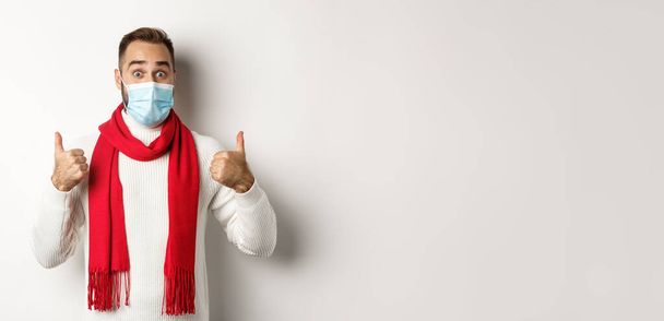 Covid-19, koncepcja zamknięcia i kwarantanny. Zaskoczony mężczyzna w masce twarzy pokazuje kciuki do góry, stojący w swetrze i czerwonym szaliku, białe tło. - Zdjęcie, obraz