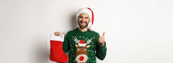 Concepto de Navidad y vacaciones de invierno. Hombre feliz y complacido en el sombrero de Santa, como regalo en calcetines de Navidad, mostrando el pulgar hacia arriba, de pie sobre fondo blanco. - Foto, imagen