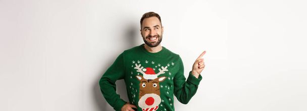 Újévi ünneplés és téli ünnepek koncepció. Jóképű mosolygós férfi zöld karácsonyi pulóver, mutatva és nézi a bal felső sarokban logó áruház, fehér háttér. - Fotó, kép