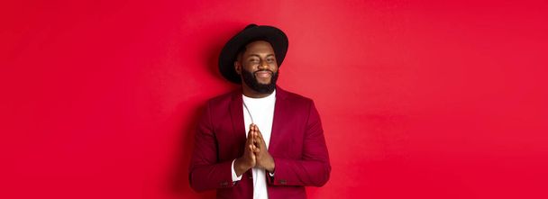 Handsome successful Black man relish profit, rub hands and smiling satisfied, standing against red background in party outfit. - Foto, Bild