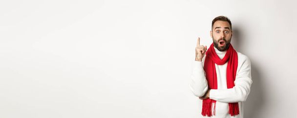 Święta Bożego Narodzenia i koncepcja świętowania. Podekscytowany brodaty mężczyzna mający pomysł, wskazujący palcem i sugerujący plan, stojący w czerwonym szaliku ze swetrem, białe tło. - Zdjęcie, obraz