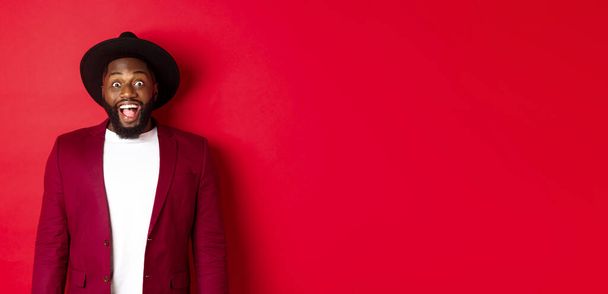 Mode und Party-Konzept. Fröhlicher afrikanisch-amerikanischer Mann in Jacke und schickem Hut, schaut erstaunt und glücklich in die Kamera, schaut sich die Neujahrspromo an, roter Hintergrund. - Foto, Bild