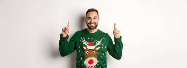 Winterurlaub und Weihnachten. Schöner bärtiger Mann in grünem Pullover, mit erhobenen Fingern lächelnd, Werbung zeigend, weißer Hintergrund. - Foto, Bild