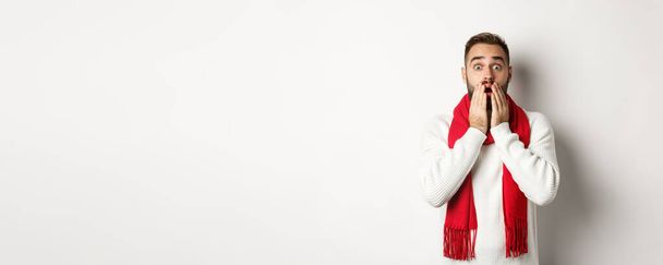 Święta Bożego Narodzenia i koncepcja świętowania. Przystojny brodaty facet w czerwonym szaliku i swetrze wyglądający na zszokowanego, pod wrażeniem reklamy, białe tło. - Zdjęcie, obraz