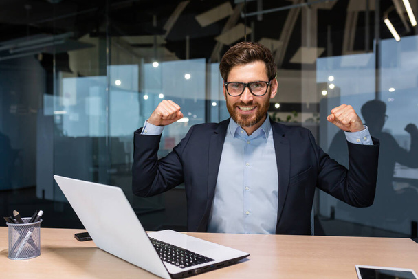 Portrait of mature senior businessman in office, man looking at camera and holding hands up super power and superhero gesture, successful investor using laptop inside building. - Zdjęcie, obraz