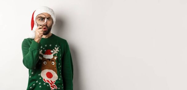 Χριστούγεννα και γιορτές έννοια. Περίεργος τύπος κοιτάζοντας κάτι μέσα από μεγεθυντικό φακό, στέκεται σε Σάντα καπέλο με πουλόβερ Χριστούγεννα, στέκεται πάνω από το λευκό φόντο. - Φωτογραφία, εικόνα