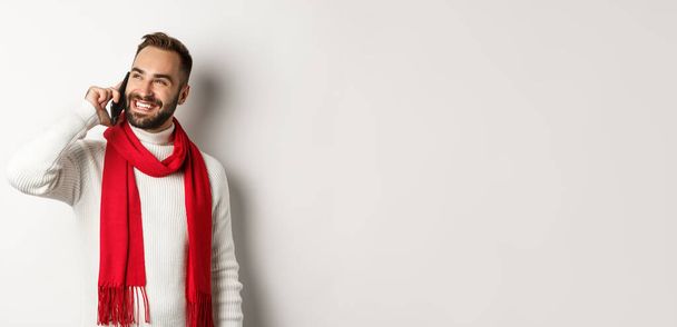 Handsome smiling man talking on phone, looking satisfied, standing in winter scarf and sweater, white background. - Fotografie, Obrázek