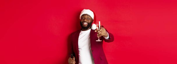 Weihnachten, Party und Feiertage Konzept. Schöner schwarzer Mann mit Weihnachtsmannhut hebt ein Glas Champagner und lächelt, spricht Toast, feiert Neujahr, roter Hintergrund. - Foto, Bild