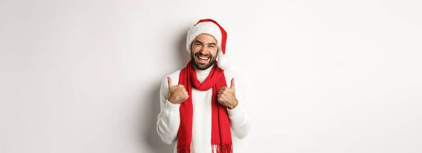 Christmas holidays. Happy man at good new year party showing thumb up, enjoying celebration, wearing santa hat and red scarf, white background. - Photo, image