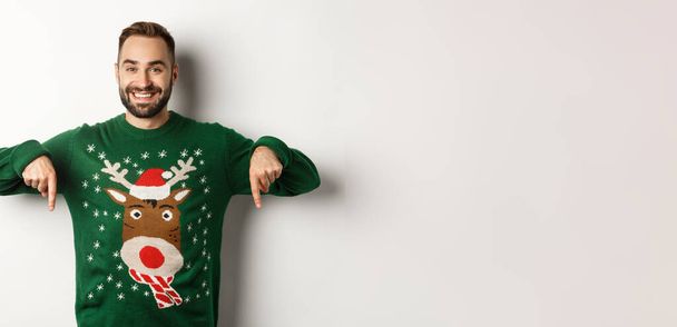 Winterurlaub und Weihnachten. Schöner lächelnder Mann mit Bart, Zeigefinger nach unten und Promoangebot, in grünem Pullover, vor weißem Hintergrund stehend. - Foto, Bild