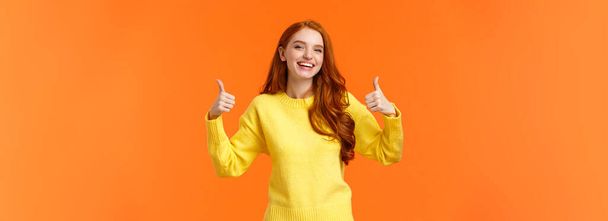 Holidays, gestures, people concept. Cheerful cute redhead girl smiling and showing thumbs-up recommend product, give positive reply, agree or like something, nod agreement, orange background. - Photo, Image