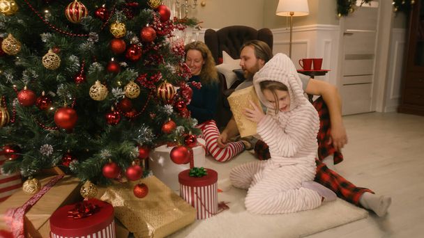 Happy caucasian family opening gifts on Christmas morning under big decorated Christmas tree. Warm atmosphere at home on Christmas or New Year. Winter holidays. Festive home decorations. - Foto, immagini