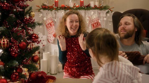 Onnellinen valkoihoinen perhe avaamassa joululahjoja koristellun joulukuusen alla. Äiti purkaa punainen juhlava kimaltava mekko. Lämmin tunnelma kotona jouluna tai uutena vuotena. Talvilomat. - Valokuva, kuva