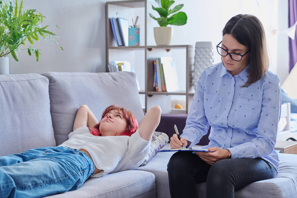Psychologist helping teenage female, individual therapy in doctors office. Professional counselor listening teenager, girl lying on couch. Mental health, adolescence, psychology, psychiatry concept - 写真・画像