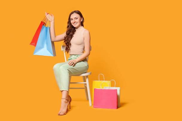Happy woman holding colorful shopping bags on chair against orange background - Фото, зображення