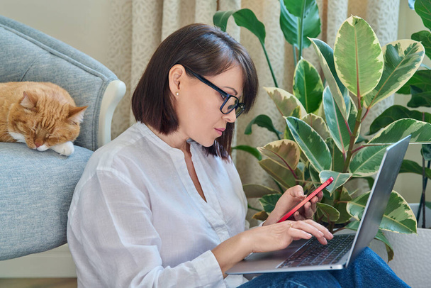 Middle aged woman sitting on floor using smartphone laptop. Female in home interior with sleeping pet ginger cat on armchair. Work at home, technology, leisure, freelancing, lifestyle, people, animal - Fotó, kép