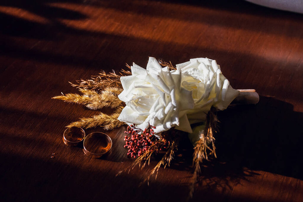 Golden wedding rings and the groom's boutonniere made of white roses and brown grass, lie on a wooden brown table. - Photo, Image
