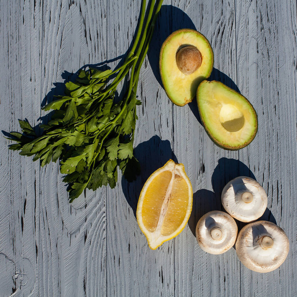 Kitchen composition of fresh avocados, lemon, raw champignons and greens, parsley on a light textured wooden table. - Photo, image
