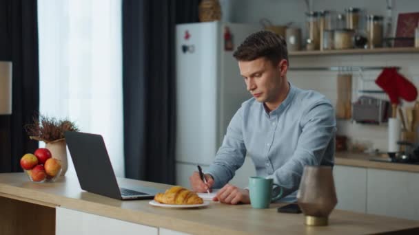 Focused man making notes working laptop in comfortable kitchen alone. Satisfied young businessman smiling checking business results on computer. Attractive freelancer using wireless technology. - Footage, Video
