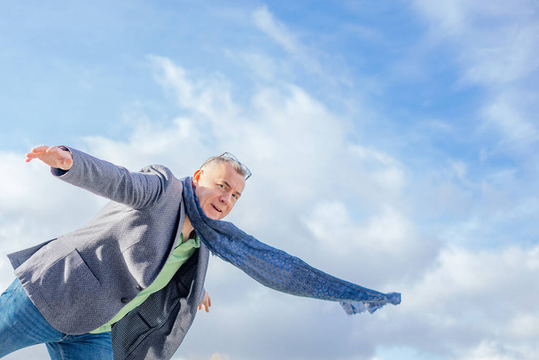 Portrait of handsome middle-aged man with short hair wearing jacket, warm blue scarf, glasses, jeans, balancing standing on one leg on cloudy sky background, stretching hands, looking at camera. - 写真・画像