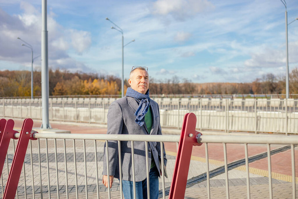 Portrait of confident middle-aged man with short grey hair wearing jacket, warm blue scarf, jeans, standing on concrete tiles square near red white railings, walking outdoors on sunny day in autumn. - Foto, imagen