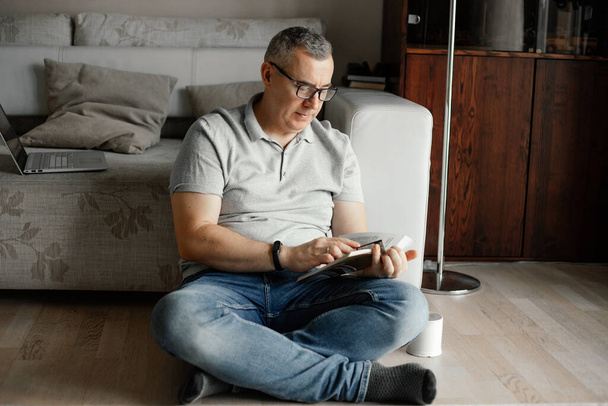 Portrait of concentrated middle-aged man with short hair wearing T-shirt, glasses, jeans, sitting with crossed legs near sofa on floor, holding reading book at home. Spending free time, education. - Foto, afbeelding