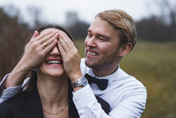 Ο άντρας καλύπτει τα μάτια της γυναίκας του με τα χέρια του. Νεαρό παιχνιδιάρικο ζευγάρι την ημέρα του γάμου τους. Υψηλής ποιότητας φωτογραφία - Φωτογραφία, εικόνα