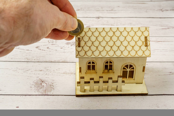 Maison en bois miniature et pièce en euros introduite à la main. Concept d'épargne pour l'achat d'une maison. Hausse des taux d'intérêt hypothécaires - Photo, image