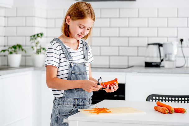 Preteen girl pilling carrot at kitchen and smiling. Child with orange vegetables food cooking at home - Zdjęcie, obraz