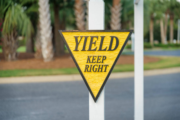 収量のある木彫りの道路標識は、フロリダ州マイアミで右を保ちます。道路や木々の景色に対する黄色と黒のフレームと文字でポストにイールドサインのクローズアップ. - 写真・画像