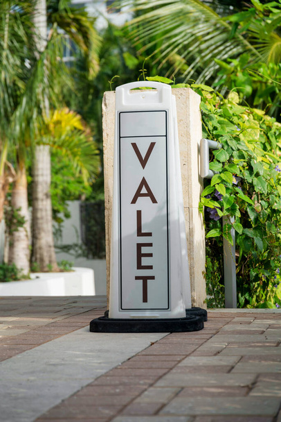 Floor-mounted Valet signage on a bricks pavement- Miami, Florida. Valet sign near the railings with crawling plants on the right and views of trees on the left. - Photo, Image