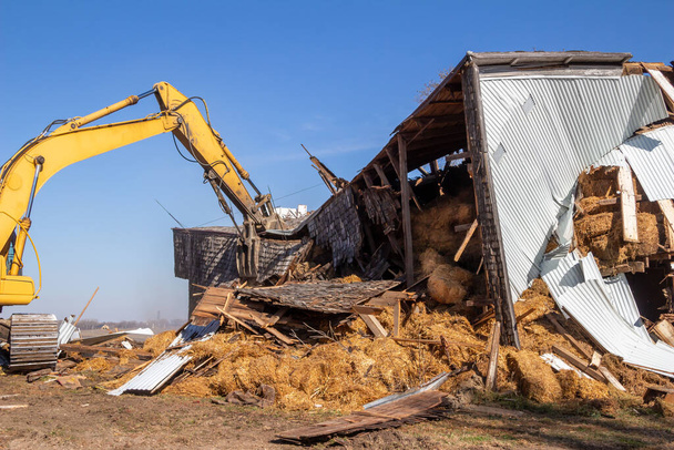 Terk edilmiş eski bir ahır binasının ağır bir ekipman kazıcısıyla yıkılışının yakın görüntüsü. - Fotoğraf, Görsel