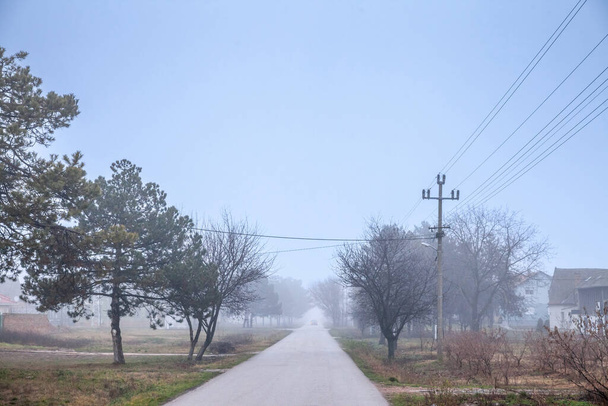 Selective blur on an empty road and street in the village of Vladimirovac in Vojvodina, Banat, Serbia, in the countryside, with a smog and fog during a cold freezing afternoon of winter. - Foto, Bild