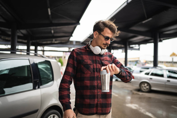 Один чоловік молодий дорослий білий чоловік з вусами, що стоять на парковці, тримає чашку чаю або кави, чекає і дивиться на наручний годинник, перевіряючи час, коли справжні люди копіюють простір
 - Фото, зображення