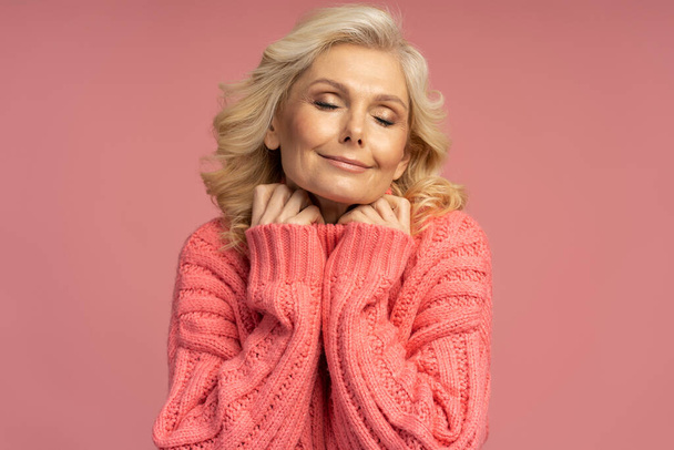 ピンクの背景に隔離されたスタイリッシュな冬のセーターを身に着けている美しい幸せな成熟した女性  - 写真・画像