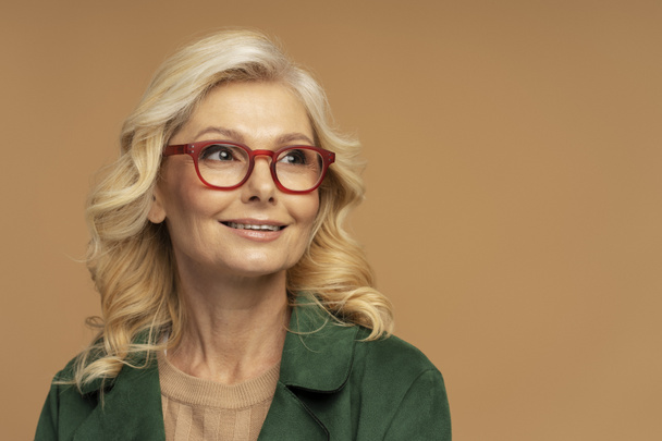 Porträt einer lächelnden reifen Frau mit roter stylischer Brille, die isoliert auf beigem Hintergrund wegschaut, Kopierraum. Vision-Konzept   - Foto, Bild