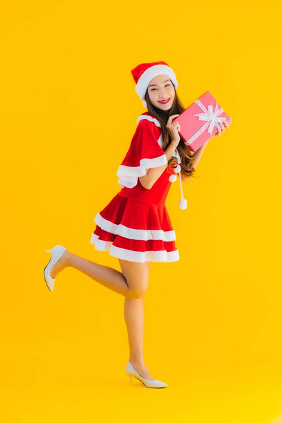 Güzel genç Asyalı noel kıyafetleri ve şapka gülümsemesi sarı izole edilmiş arka planda kırmızı hediye kutusuyla mutludur. - Fotoğraf, Görsel