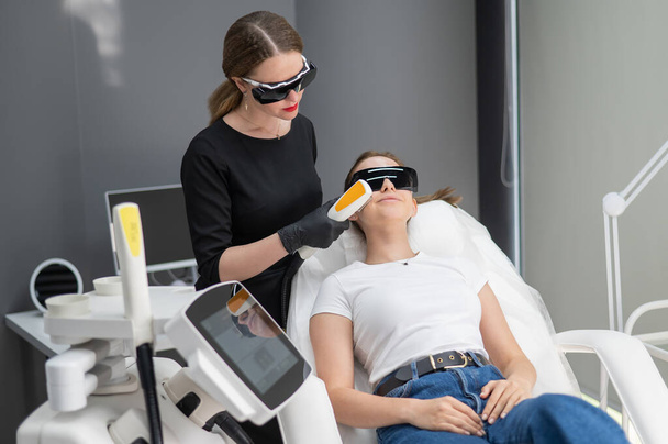 Mujer caucásica joven con gafas en el procedimiento de fotorejuvenecimiento. Médico cosmetólogo con una paciente femenina - Foto, Imagen