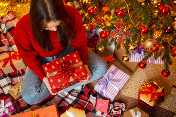 Πάνω-κάτω πλάνο ενός κοριτσιού που κάθεται κάτω από διακοσμημένο χριστουγεννιάτικο δέντρο με ένα δώρο. Χριστουγεννιάτικη γιορτή - Φωτογραφία, εικόνα
